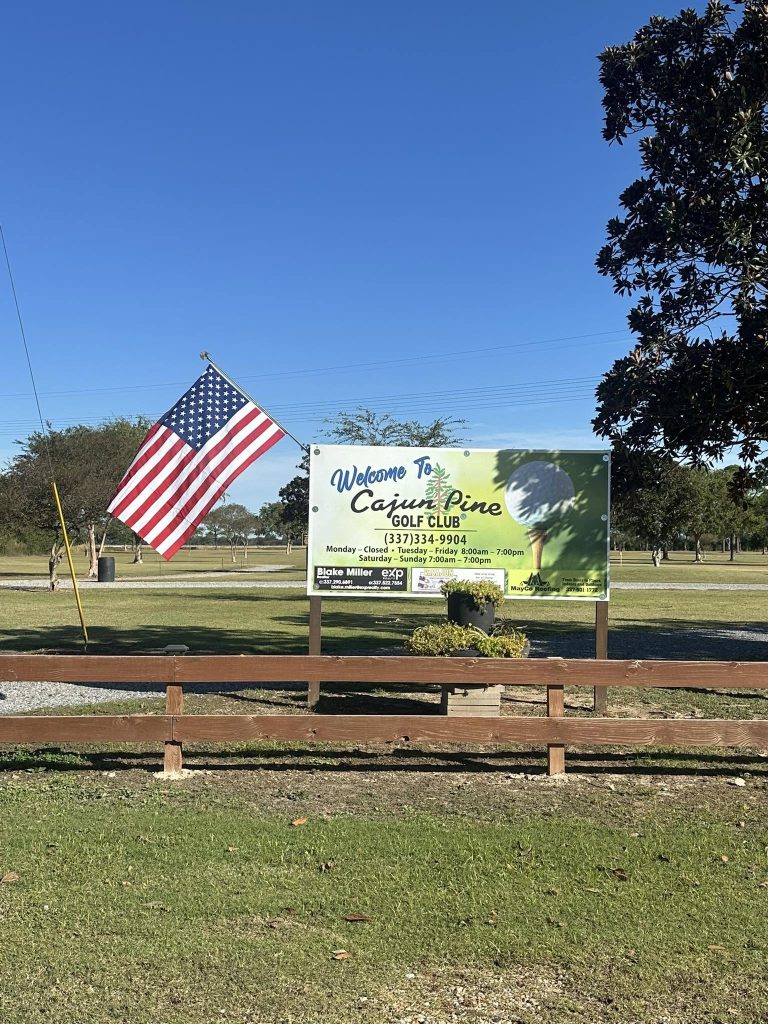 Large sign that reads Cajun Pine Golf Club with an American Flag in front of a golf course